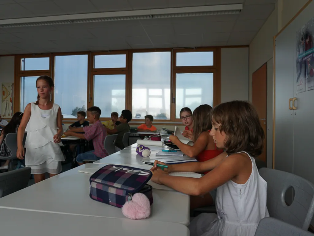 Lehrkraft steht an der Tafel, Kinder arbeiten an den Tischen.